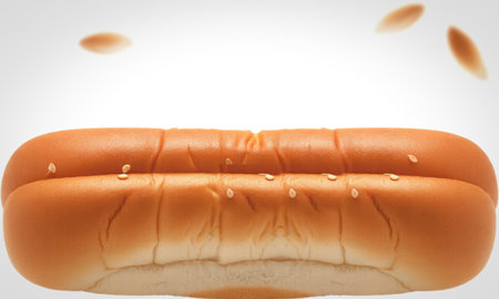 A close-up of a freshly baked hot dog bun with sesame seeds sprinkled on top. Several sesame seeds are captured mid-air, falling towards the bun.の写真素材