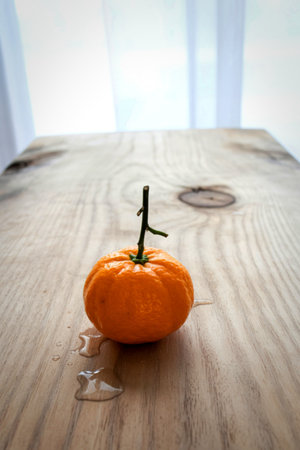 A solitary, bright orange fruit with a thin stem sits centered on a textured wooden table, with soft light in the background.の写真素材