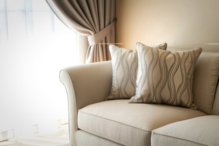 A close-up view of a plush beige sofa featuring two textured decorative pillows and soft, flowing drapes near a window.の写真素材