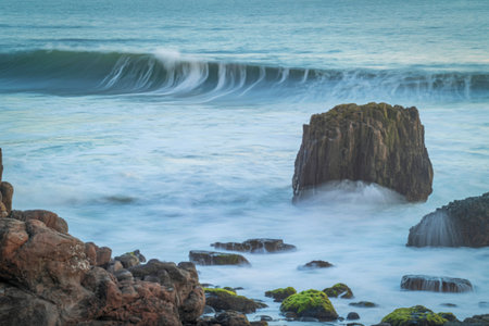 Powerful ocean waves surge and break against a large, solitary rock formation and the rocky shoreline, creating a dynamic seascape.の写真素材