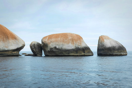 Several massive, weathered granite boulders rise from the tranquil, blue-grey ocean under a cloudy sky.の写真素材