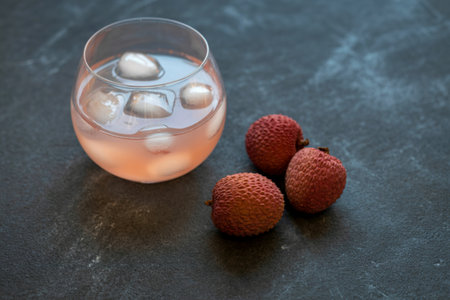 A clear glass filled with a pale pink beverage and ice cubes sits next to three ripe lychee fruits on a dark, textured background.の写真素材