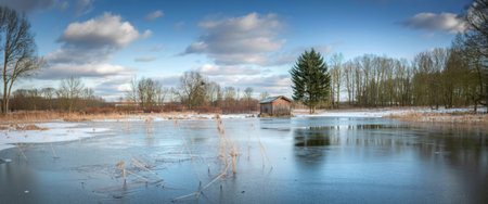 A serene winter scene featuring a partially frozen pond reflecting the sky, surrounded by bare trees and a small building.の写真素材