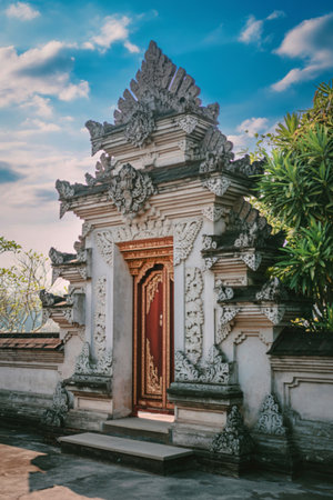 A detailed, ancient stone temple gate with intricate carvings and a wooden door, set against a backdrop of lush greenery and a cloudy sky.の写真素材