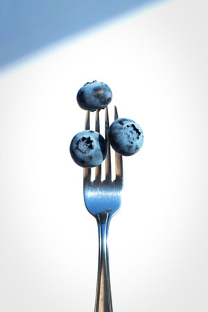 A close-up shot of three plump, dark blue blueberries impaled on the tines of a shiny metal fork against a bright white background.の写真素材