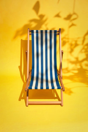 A classic striped deck chair sits on a vibrant yellow background, casting a shadow from overhead foliage, evoking a sunny, relaxed atmosphere.の写真素材