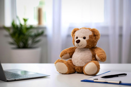 A fluffy teddy bear sits patiently at a desk next to an open laptop, pen, and notebook, with a plant in the background.の写真素材