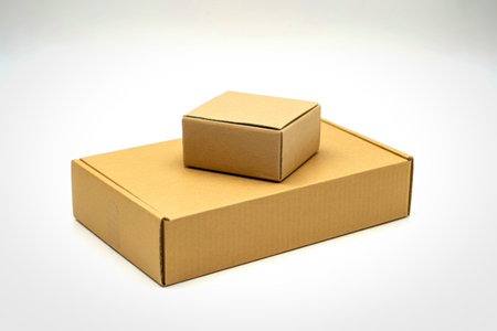 Two plain brown cardboard boxes, one larger and rectangular, the other smaller and square, are stacked on a clean white surface.の写真素材