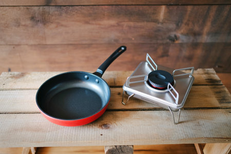 A small frying pan with a red rim sits next to a portable camping stove on a rustic wooden background.の写真素材
