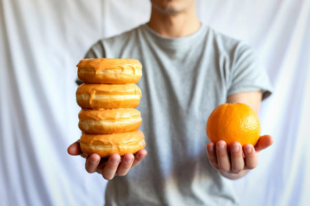 A person in a grey t-shirt holds a stack of five glazed donuts in their left hand and a bright orange in their right hand.の写真素材