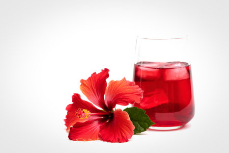 A tall glass filled with a vibrant red beverage, ice cubes, and a beautiful red hibiscus flower with green leaves beside it.の写真素材