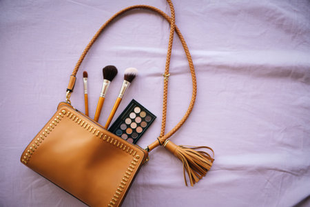 A tan leather pouch with a tassel sits open, revealing makeup brushes and an eye shadow palette, laid out on a white textured surface.の写真素材