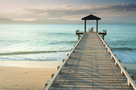 A weathered wooden pier extends over calm turquoise ocean water towards a small covered gazebo, with a sandy beach on the left.の写真素材