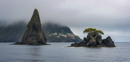 Two dramatic rock formations emerge from the calm sea, with a village nestled on a distant, fog-shrouded hillside.の写真素材