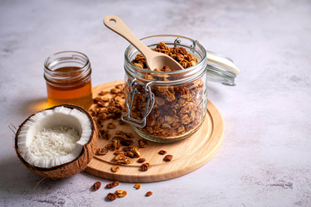 A jar of crunchy granola with a spoon, a half coconut, and a small jar of honey are artfully arranged on a light-colored surface.の写真素材