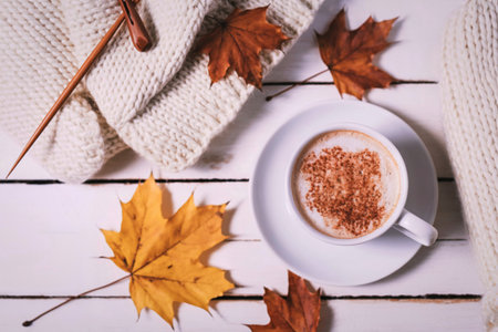 A warm cup of coffee sits on a saucer next to a soft blanket, surrounded by colorful fallen autumn leaves.の写真素材