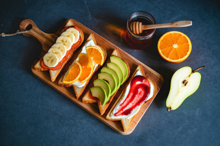 A vibrant arrangement of open-faced sandwiches featuring fruit, avocado, and jam, served with honey and fresh citrus on a rustic wooden board.の写真素材
