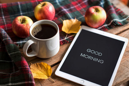 A warm mug of coffee sits beside fresh apples and autumn leaves, with a tablet screen displaying a cheerful 'Good Morning' message.の写真素材