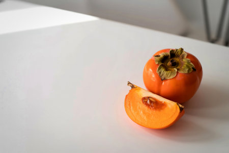 A whole, ripe persimmon sits next to a sliced portion of the same fruit on a clean white surface, with soft light illuminating the scene.の写真素材