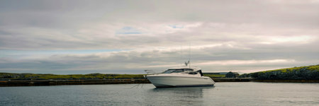 A white motor yacht rests on tranquil water, framed by a cloudy sky and lush green shoreline.