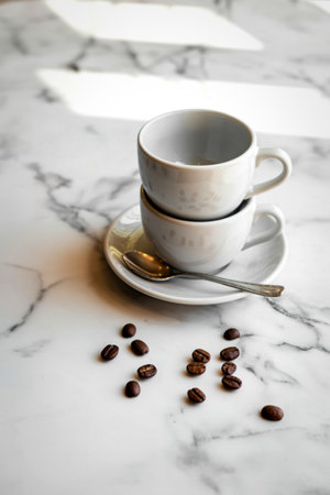 A still life featuring two stacked white coffee cups and saucer with a spoon, alongside scattered...