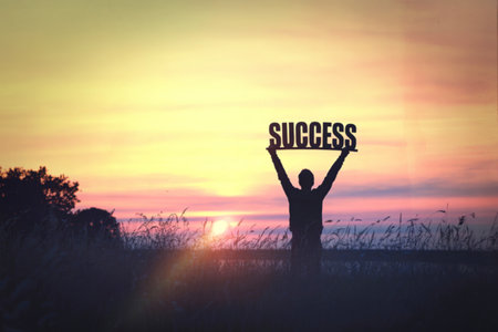 A person stands with arms raised, holding the word SUCCESS against a vibrant sunset, symbolizing...