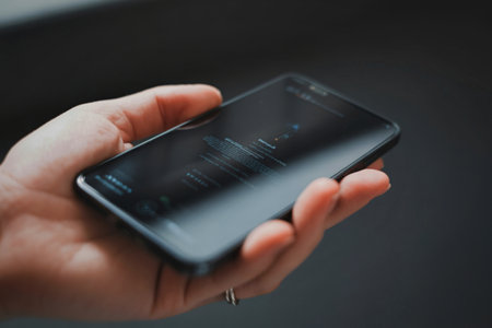 A close-up shot of a person's hand holding a dark-colored smartphone. The background is blurred and dark.の写真素材