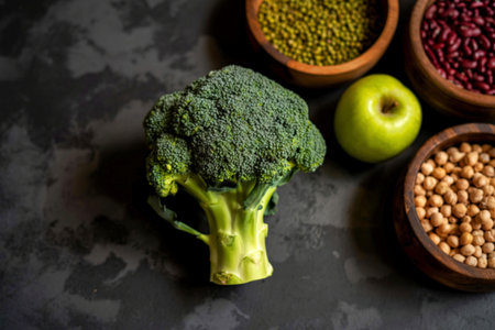 A vibrant head of broccoli sits beside a green apple and bowls of assorted beans and chickpeas, showcasing a healthy selection of ingredients.の素材