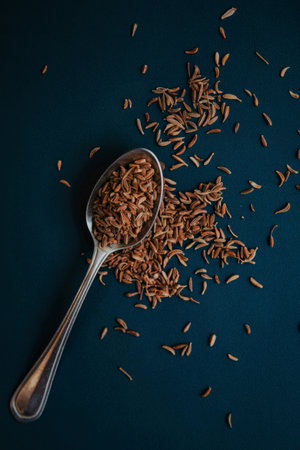 A close-up image of cumin seeds scattered on a dark blue background with a spoon holding a portion...