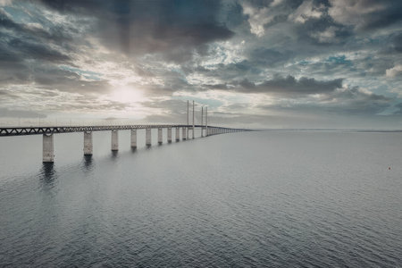 A long bridge stretches over a body of water under a cloudy sky with the sun peeking through the...
