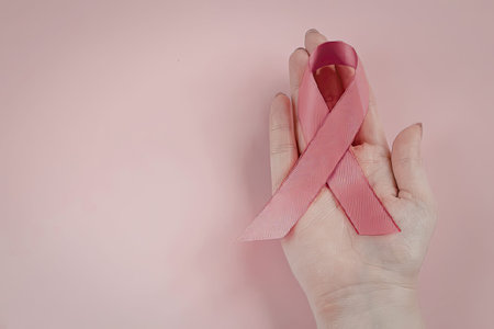 A hand holding a pink ribbon on a pink background, symbolizing breast cancer awareness