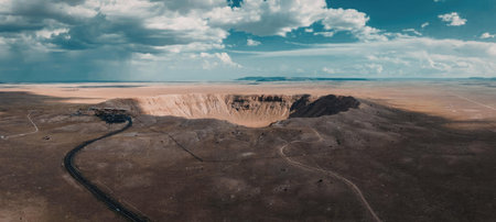 A panoramic view of a desert landscape with a vast expanse of brown terrain, a winding road, and a large cliff in the distance under a blue sky with white clouds.の写真素材