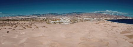 A panoramic view of a desert landscape with sandy dunes, a body of water, and a mountain range in the background under a clear blue sky.の写真素材