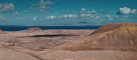 A panoramic view of a desert landscape with brown hills, a blue sea, and a blue sky with clouds.