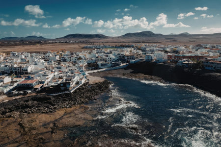 A small coastal town with white buildings and a rocky shoreline, surrounded by mountains and a blue...