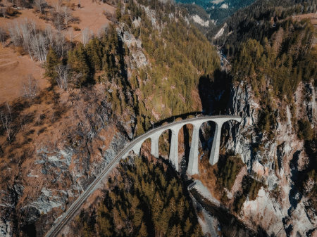 An aerial view of a bridge surrounded by mountains and trees, with a road leading to it.