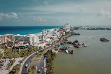 An aerial view of a coastal city with a mix of buildings, roads, and a body of water. The image showcases a scenic view of the city's waterfront, with a large body of water on the right and the ocean on the left. The city has a variety of white and brown buildings, some of which appear to be hotels or resorts. A road runs along the left side of the image, with several cars visible. The ocean is a bright blue color, while the body of water on the right is a greenish-brown color.