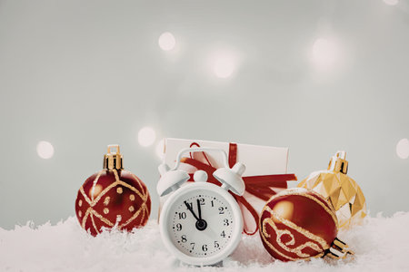 a white analog alarm clock surrounded by red and gold christmas ornaments on a snowy surface with...