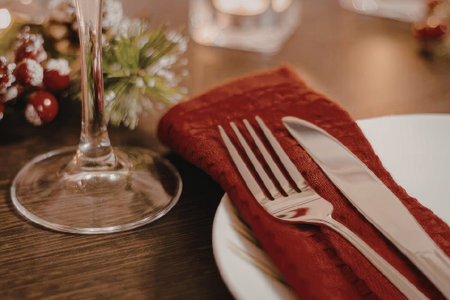 A festive Christmas dinner table setting with a red napkin, silverware, and wine glass on a wooden...
