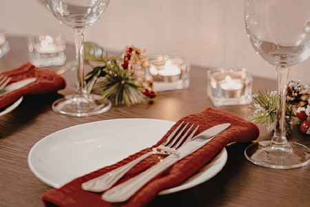 A festive Christmas dinner table setting with red napkins, silverware, and wine glasses, decorated with pine branches and candles.の写真素材