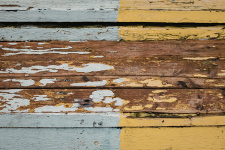 A close-up view of weathered wooden planks with peeling white, yellow, and brown paint, showcasing...