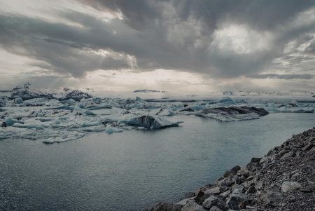 A serene yet dramatic landscape of glacier pieces floating on icy waters under a stormy sky,...