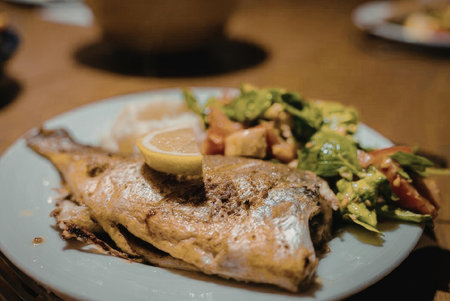 A plate of grilled fish served with a fresh salad and a slice of lemon on top, accompanied by a side...