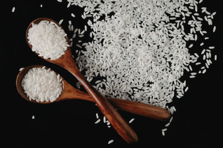 Two wooden spoons filled with white rice placed on a black background with a pile of rice beside...