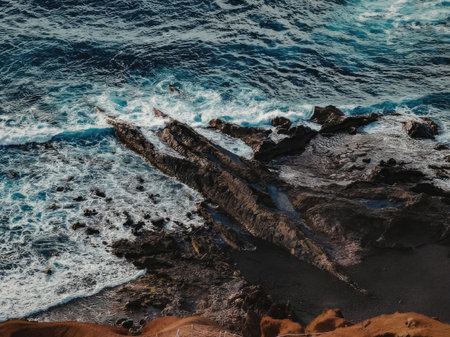 A scenic view of ocean waves crashing against a rocky shoreline, with dark rocks and red sand.
