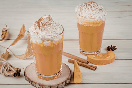 Two glasses of pumpkin spice drinks topped with whipped cream and cinnamon, accompanied by cinnamon sticks, star anise, and pumpkin slices on a wooden table.の写真素材