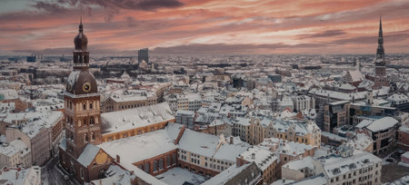 A snow-covered cityscape with a church steeple and buildings under a vibrant orange and pink sunset...