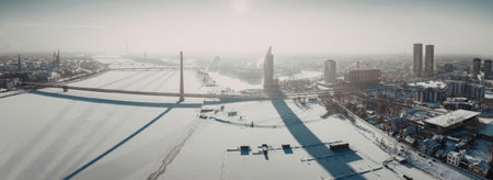 Aerial view of a snow-covered city with a bridge and river, surrounded by buildings and roads.