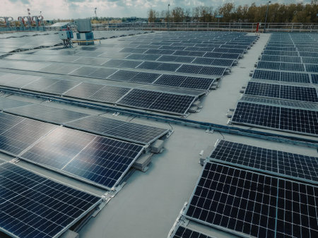 A large array of solar panels installed on a flat rooftop, with trees and a cloudy sky in the...