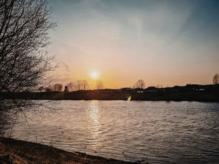 A peaceful lake scene at sunset, with the sun setting behind trees on the horizon, casting a warm...
